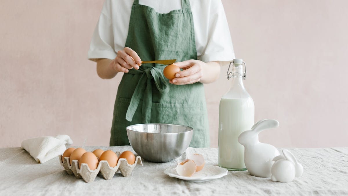 Crop anonymous female with egg in hand at table with iron bowl and glass bottle of milk